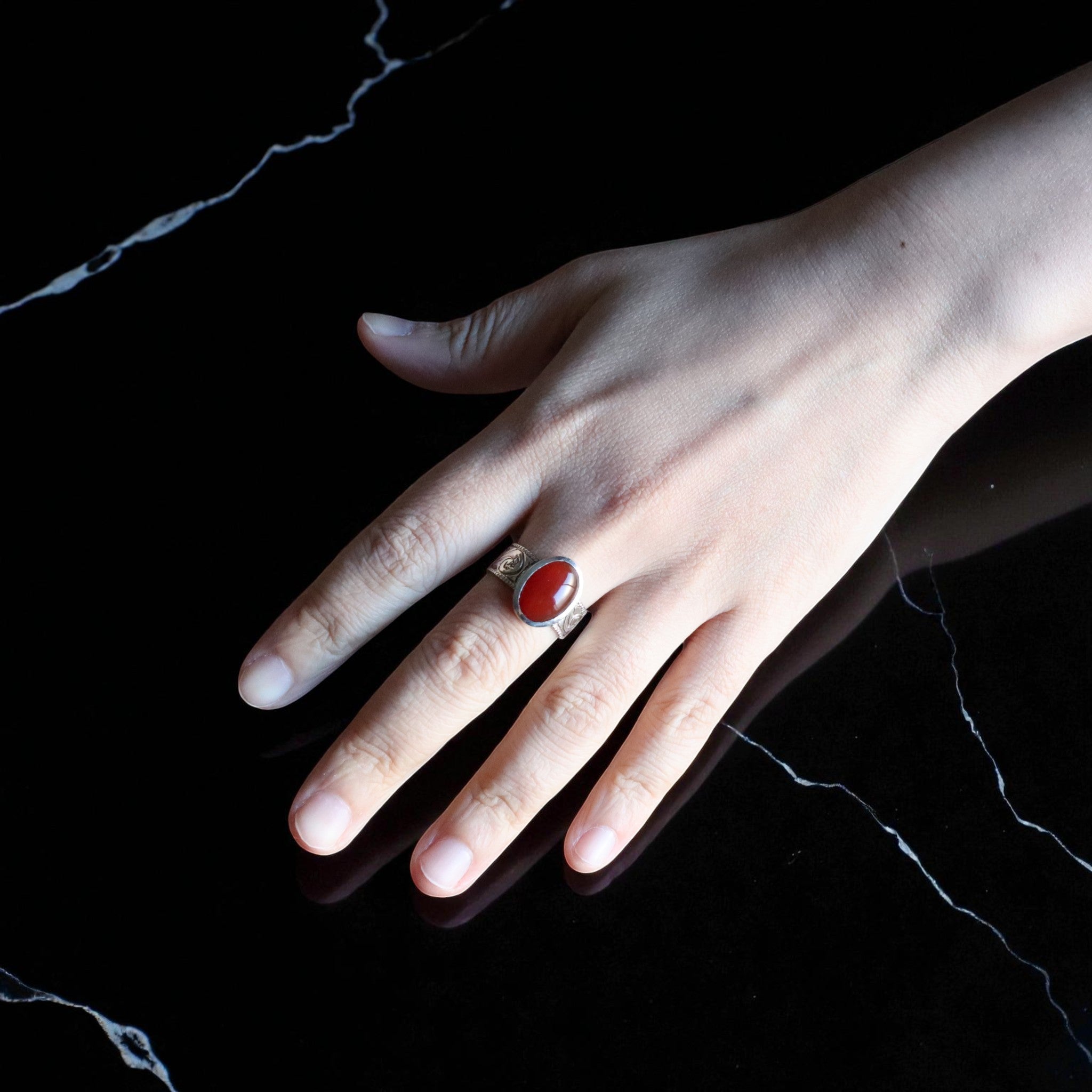 黒い大理石調の背景の上に手を置き、深紅のカーネリアンを留めたシルバーリングを着用している。石の艶やかさと金属の質感が光を受けて際立っている。