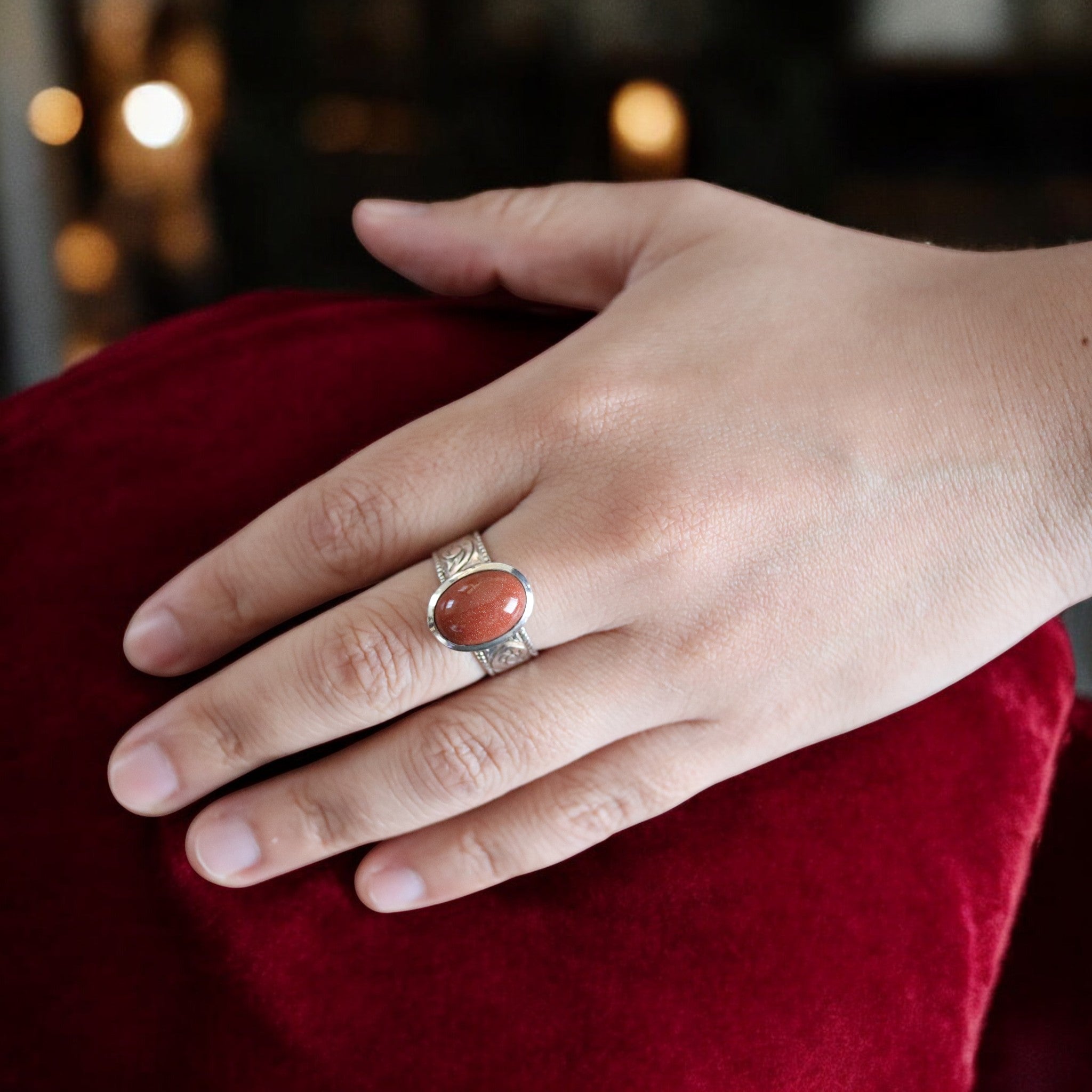 赤みを帯びたゴールドストーンを中央に留めたシルバーリングを着用した手元。石の表面に微細な光がきらめき、彫刻模様のあるリングが赤い背景の上で際立っている。