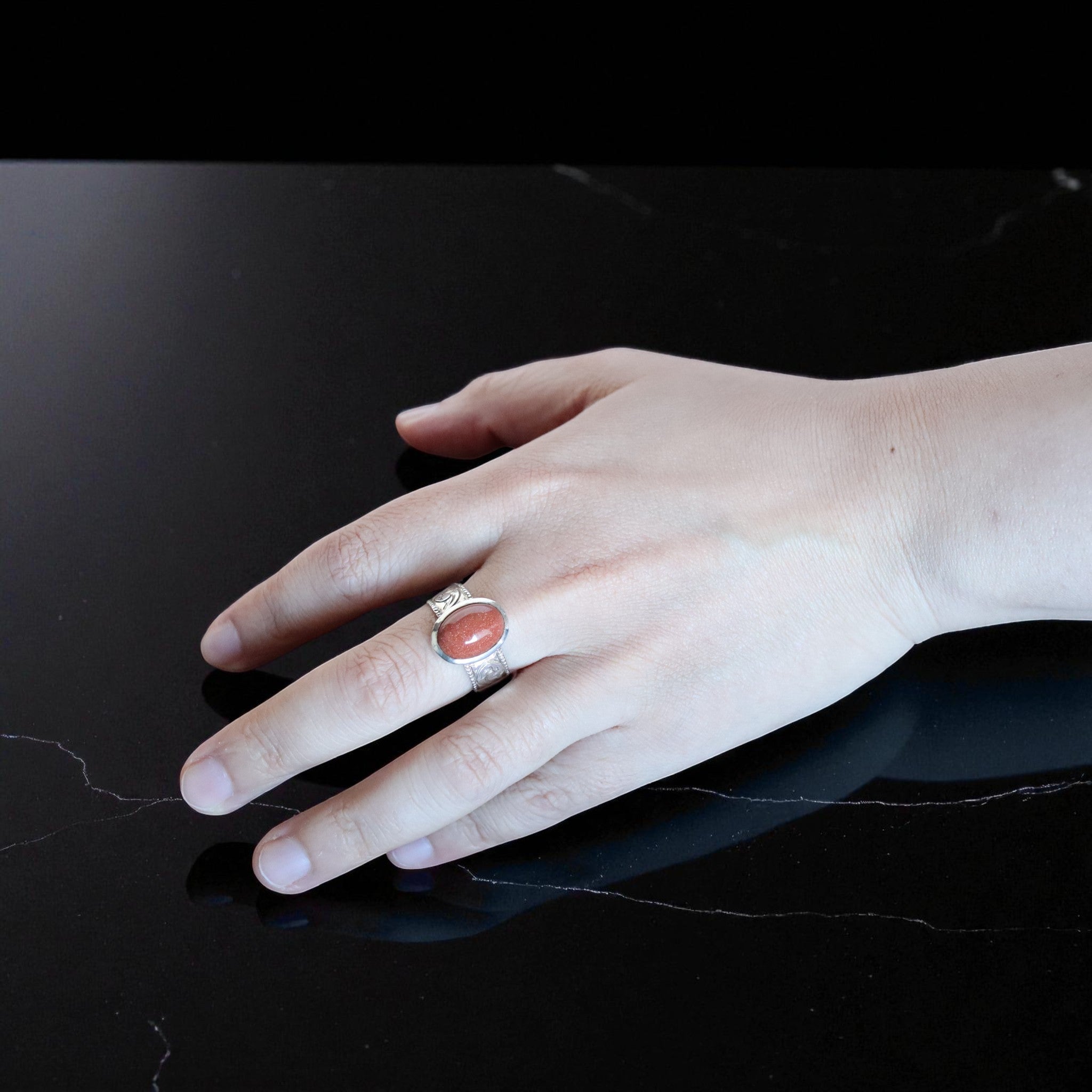 黒い大理石調の背景の上に手を置き、赤みを帯びたゴールドストーンを留めたシルバーリングを着用している。石の表面に微細な光が反射し、金属の彫刻模様が繊細に浮かび上がっている。